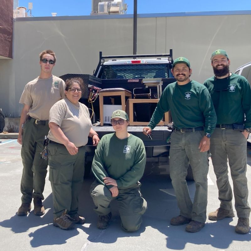 A group of five park rangers stand together smiling in a sunny parking lot in front of a pickup truck loaded with Surplus items. Two wear tan shirts and three wear green shirts with matching green caps.
