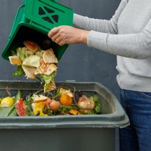 woman dumping food scraps out of a bucket into a green compost bin