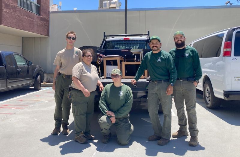 A group of five park rangers stand together smiling in a sunny parking lot in front of a pickup truck loaded with Surplus items. Two wear tan shirts and three wear green shirts with matching green caps.