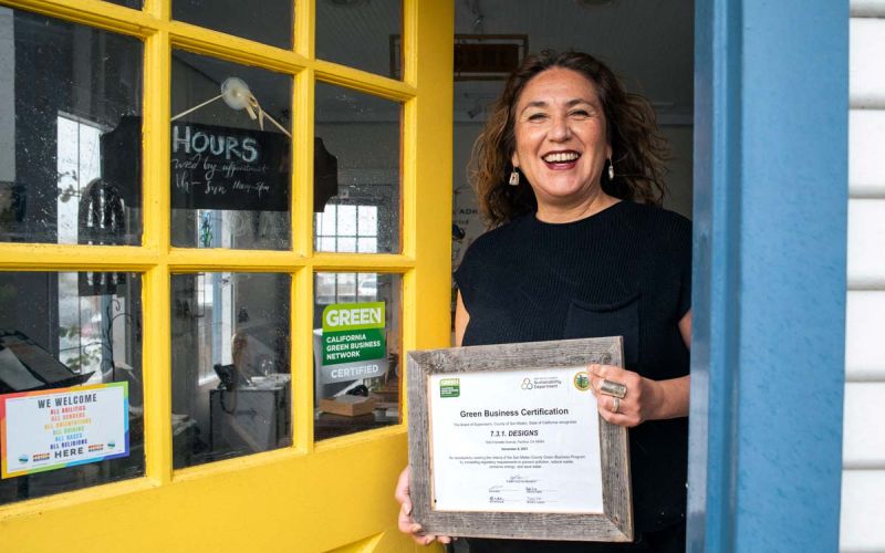 woman owner of 7.3.1. Designs in Pacifica holds framed Green Business Certification award in business doorway