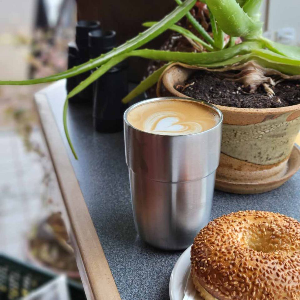 Okapi reusable cup filled with a latte next to a bagel at a cafe
