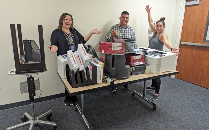 three individuals pose behind a table of surplus materials including printers and printer paper