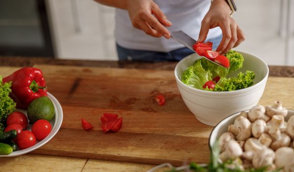 A person adds sliced tomatoes to a salad bowl on a cutting board