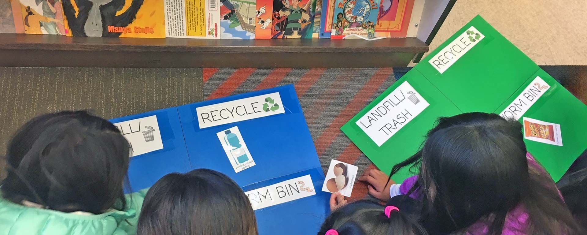 elementary students playing waste reduction games on school library floor next to bookcase