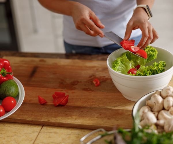 A person adds sliced tomatoes to a salad bowl on a cutting board