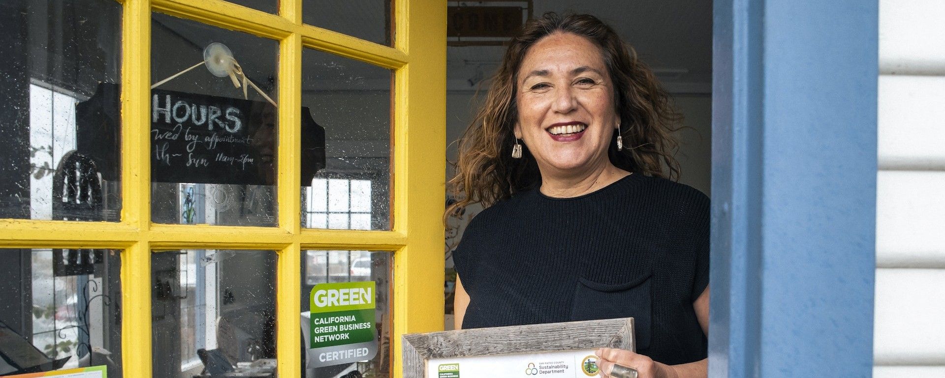 A woman stands at a business entrance holding up a Green Business certificate.