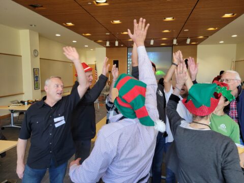 Image of many people raising their hands to high five in the middle. Three people are wearing green and red festive hats. 