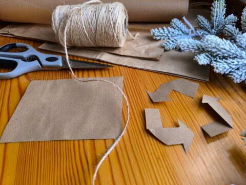 Scissors, brown paper, twine, and a tree branch on a wood tabletop.
