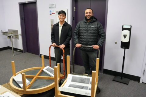 Image of two men smiling. They are holding two tables and keyboards on rolling dollies. 