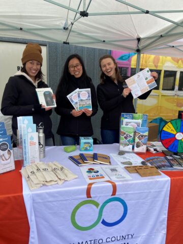 Three Sustainability Department staff at a tabling booth holding up branded materials.