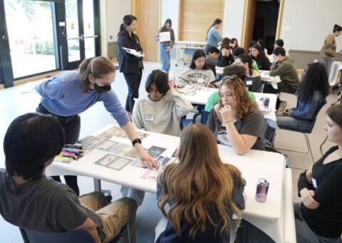 5 students sitting around a table with a climate education board game. A mentor is explaining the game to them.